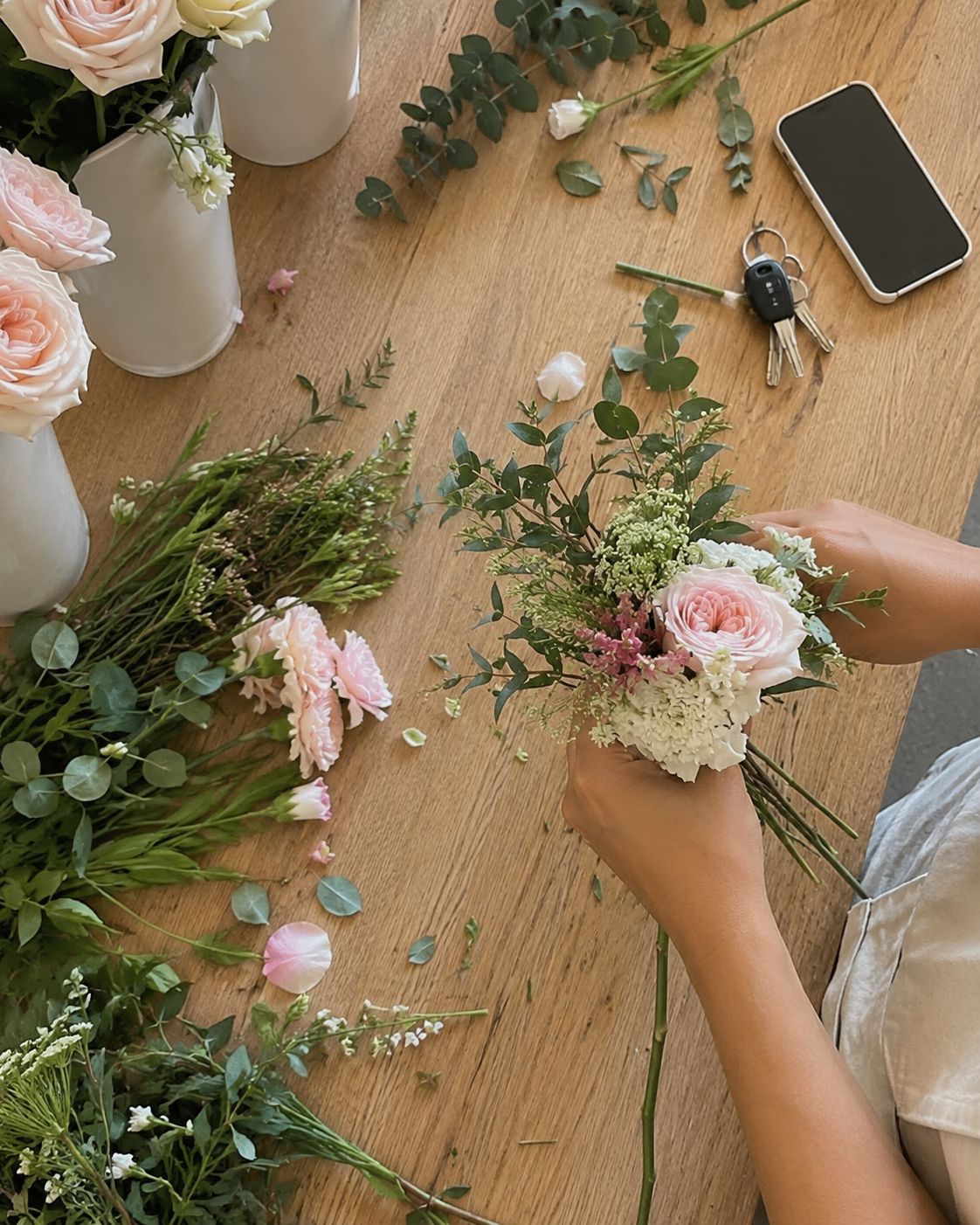 Overhead view of hands gathering roses, carnations and eucalyptus into a small bouquet on a timber table